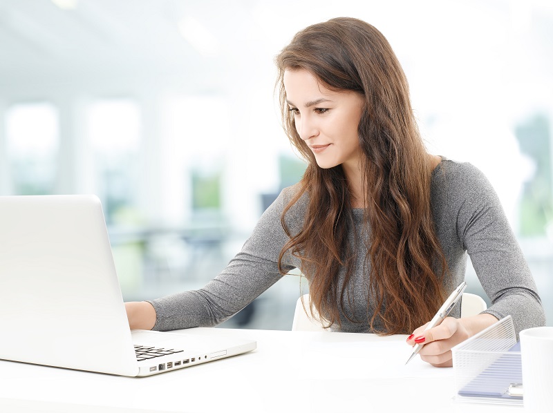 Eine junge Frau in einem hellen Büro sitzt an einem Tisch und benutzt ihren Laptop, während sie sich zeitgleich Notizen macht.