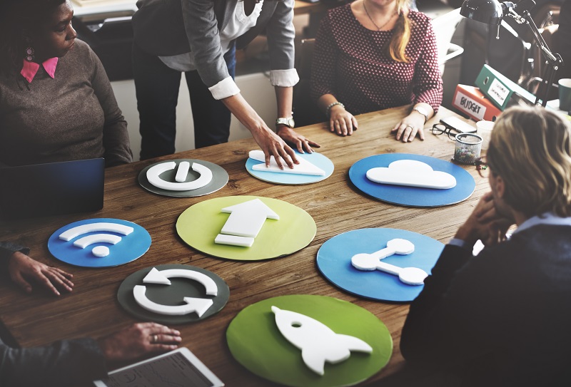 Eine Gruppe von Mitarbeitern steht diskutierend an einem Tisch, auf dem verschiedene ikonische Symbole als Plastikformen ausgelegt sind, unter anderem die Symbole für WLAN, Cloud, Seite neu laden und Teilen per Social Media.