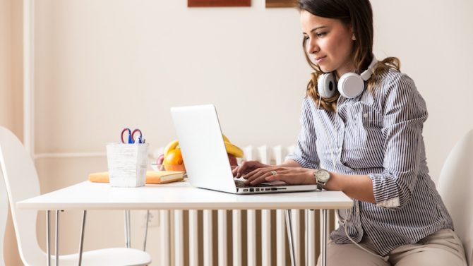 Eine junge Frau mit Kopfhörern sitzt an einem Küchentisch, auf dem sich eine Obstschale, ein Buch und Stifte befinden, und arbeitet mit ihrem Laptop.