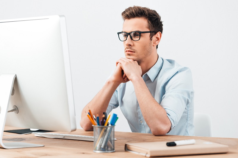 Ein junger Mann mit Brille sitzt an einem Schreibtisch und betrachtet den Bildschirm eines Arbeits-PCs.