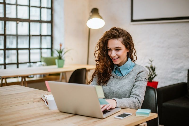 Eine junge Frau sitzt mit einem Buch, einer Tasse Kaffee, einem Stapel Post-Its und ihrem Laptop an einem Tisch, um sich über die Indesign-Grundlagen zu informieren.