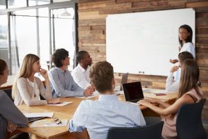 Eine Frau in einem Konferenzraum erklärt dem versammelten Team an einem Whiteboard die Indesign-Grundlagen.