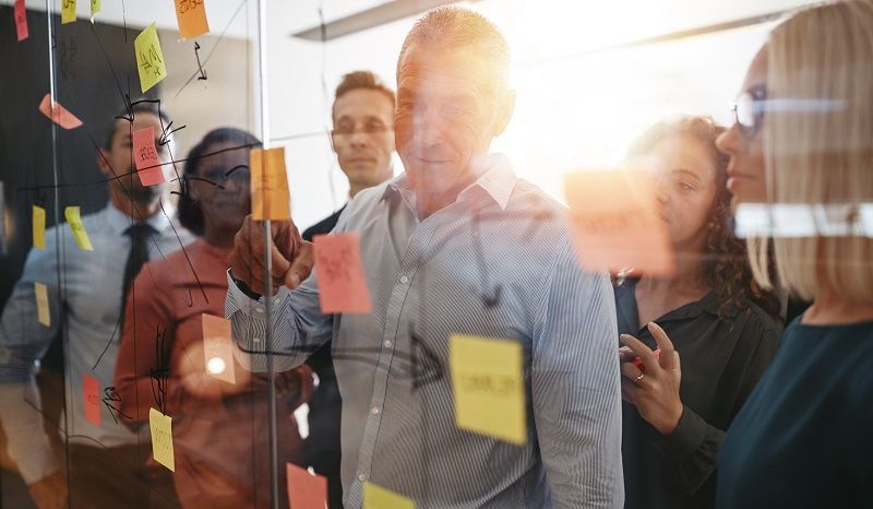 Eine Gruppe von Mitarbeitern bei der Durchsicht von Notizen zum Workflow-Management an einer gläsernen Tafel.