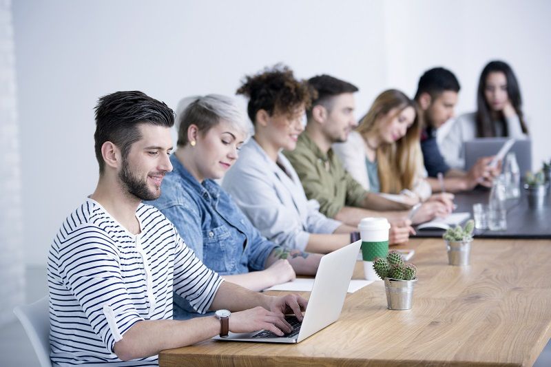 Eine Gruppe von Mitarbeitern sitzt an einem Tisch, ein Teammitglied greift mit dem Laptop auf ein Markenportal zu.